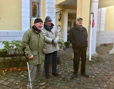 Et billede af tre personer, der står udenfor en gul bygning med hvide søjler ved indgangspartiet.